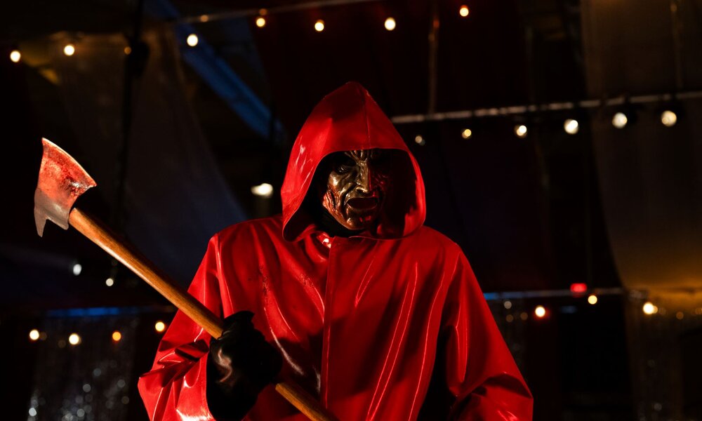 Gruselige Gestalt mit verzerrter Maske, rotem Cape und Axt steht dramatisch beleuchtet unter bunten Lichterketten im Dunkeln. | © Alan Markfield/Netflix 