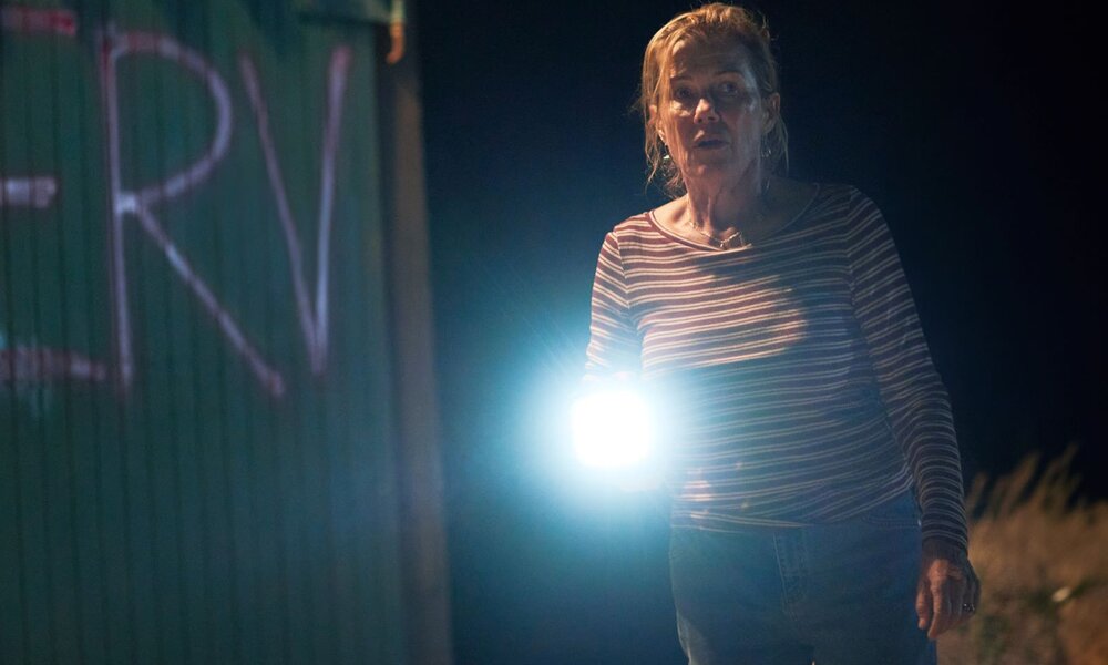 Bei Nacht leuchtet eine ältere, besorgte Frau mit zerzaustem Haar und Taschenlampe vor bemalter grüner Wand in die Kamera. | © Netflix