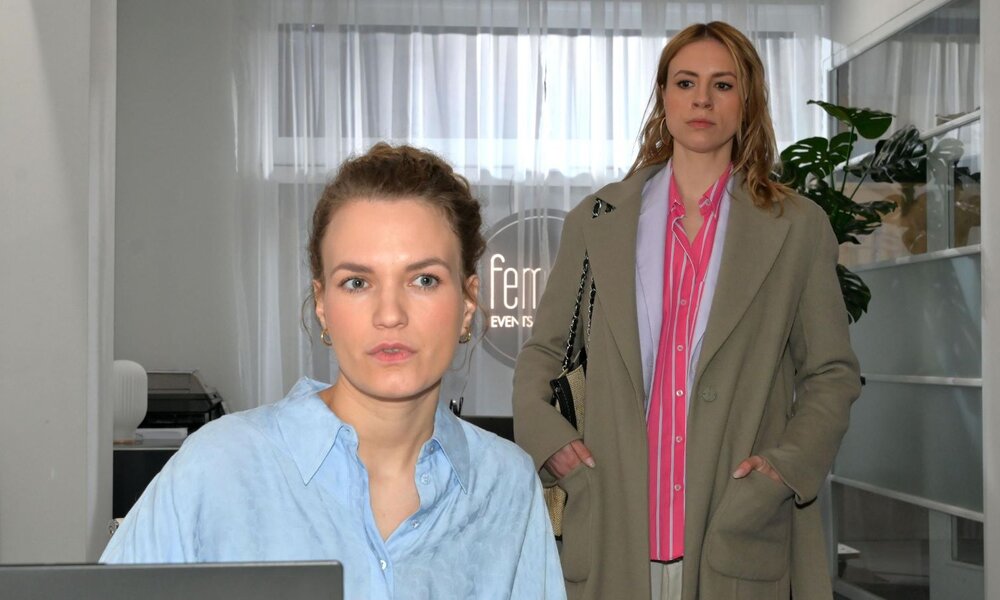 Junge Frau mit Dutt in hellblauer Bluse sitzt konzentriert am Schreibtisch, im Hintergrund betritt eine ernste Frau das Büro. | © RTL / Rolf Baumgartner