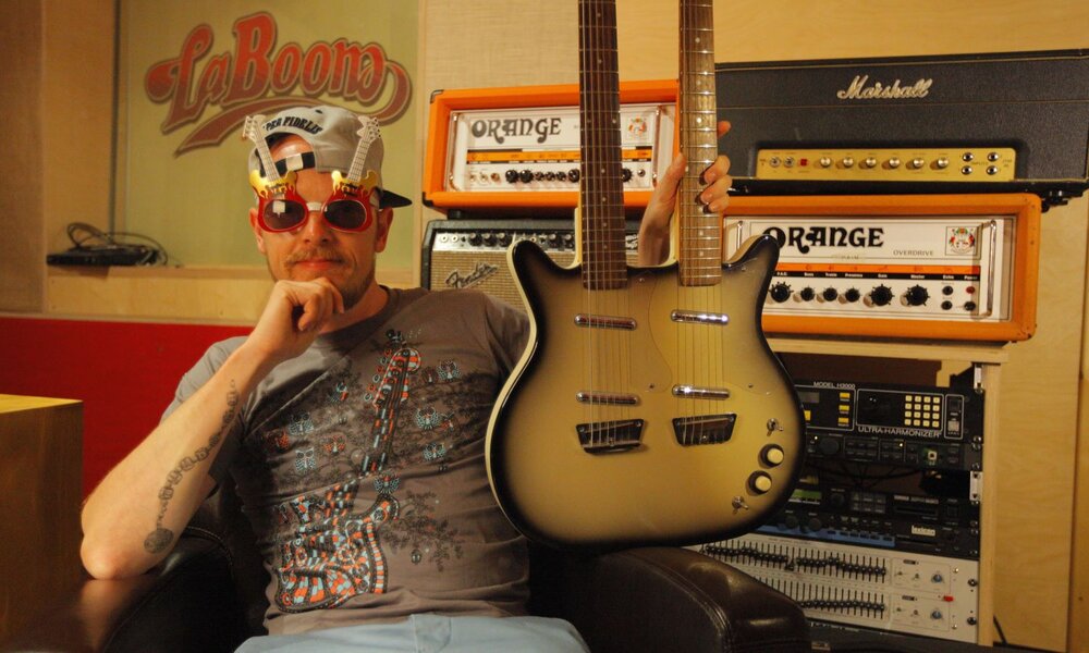 Ein Mann mit schräger Sonnenbrille und Cap hält eine zweihalsige E-Gitarre, umgeben von Orange- und Marshall-Verstärkern. | © SWR/Vibrophil Media, 2024