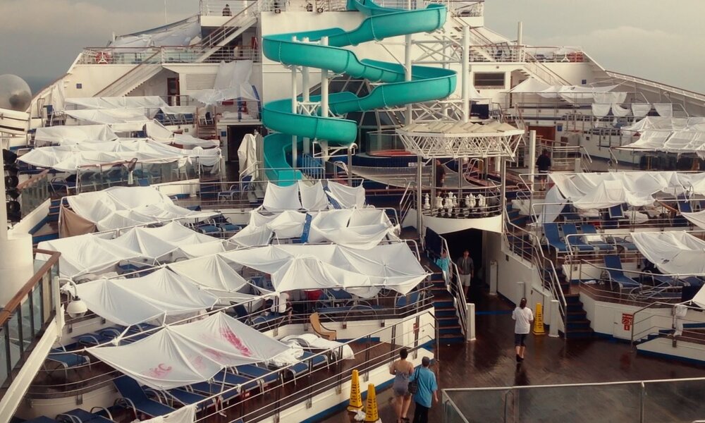 Blick auf das Deck eines Kreuzfahrtschiffs, auf dem Liegen mit weißen Laken abgedeckt sind, im Hintergrund eine grüne Wasserrutsche. | © Netflix