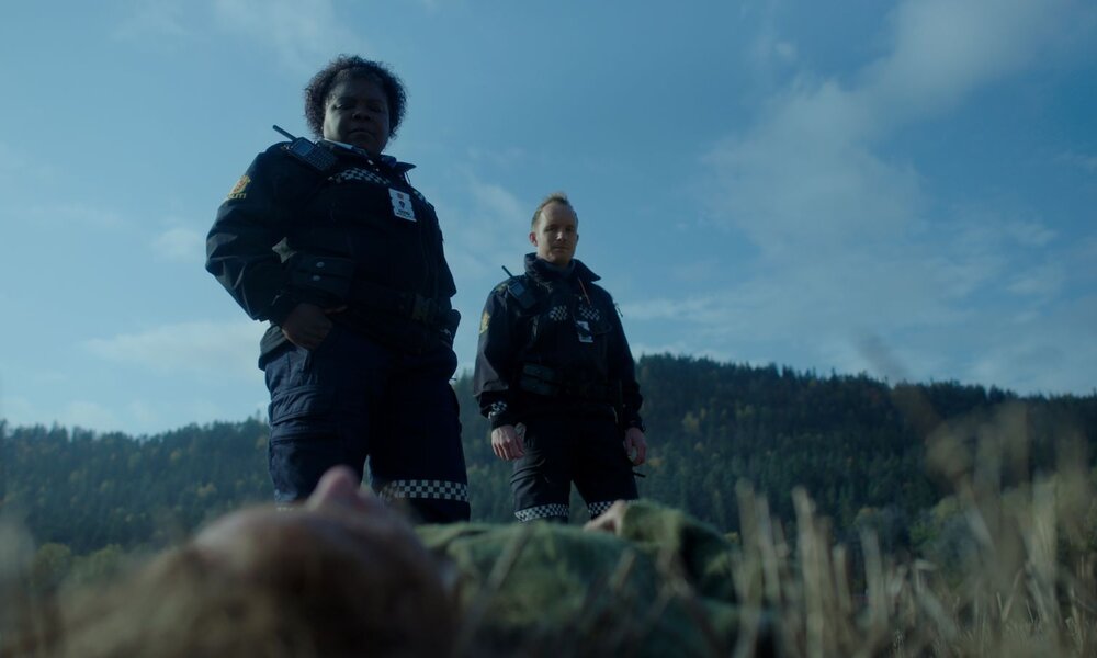Szene aus "Post Mortem": Zwei Polizist:innen mittleren Alters blicken ernst auf eine leblose Person im Gras vor bewaldetem Hügel und blauem Himmel. | © Netflix