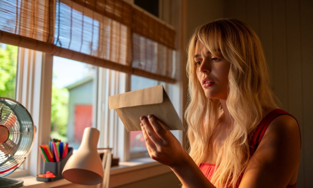Junge blonde Frau in rotem Top betrachtet nachdenklich einen Briefumschlag im warmen Licht eines Schreibtisches am Fenster. | © Amazon Content Services LLC