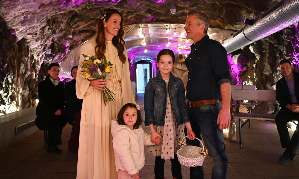 Eine Braut im cremefarbenen Kleid hält den Blumenstrauß und lächelt zwei Mädchen und einen Mann in Jeans in einer Höhlenkapelle an. | © RTL / Julia Feldhagen