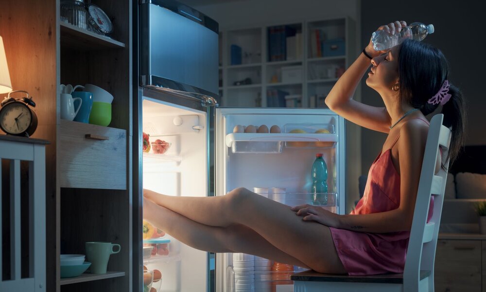 Junge Frau in rosa Satin-Nachthemd sitzt nachts mit Beinen im offenen Kühlschrank und kühlt sich mit einer Wasserflasche die Stirn. | © StockPhotoPro/stock.adobe.com