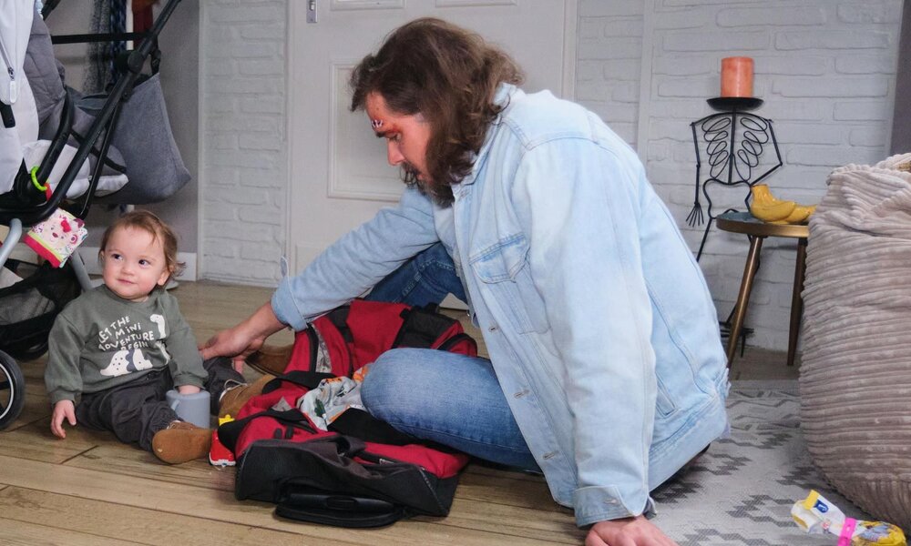Kleines Kind mit braunen Locken sitzt spielend neben einem Mann in Jeansjacke vor einem Kinderwagen in heller Wohnung. | © RTL / Stefan Behrens