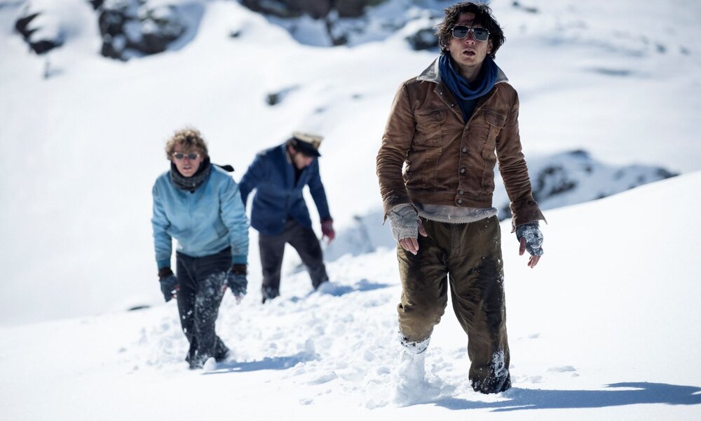 Drei erschöpfte Männer in Winterkleidung stapfen durch tiefen Schnee auf einem vereisten Berghang bei grellem Sonnenlicht. | © Netflix