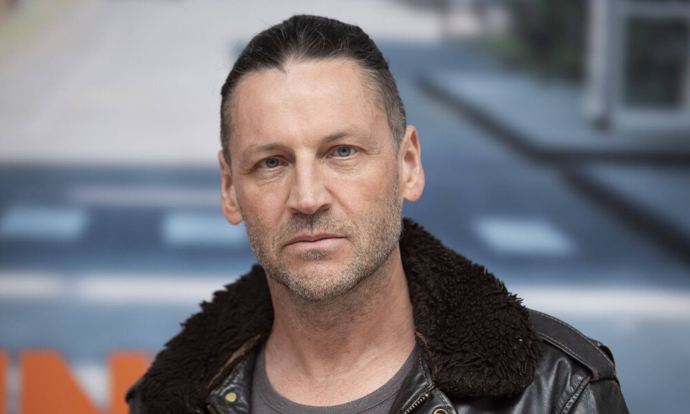 Ein ernster, mittelalter Mann mit zurückgebundenen dunklen Haaren trägt eine Lederjacke mit Fellkragen vor unscharfem Stadtmotiv. | © IMAGO / Sven Simon