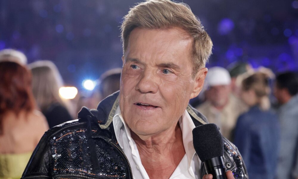 Älterer Mann mit gegeltem blonden Haar spricht mit ernstem Blick ins Mikrofon, trägt Glitzerjacke auf nächtlicher Bühne. | © IMAGO / R. Schmiegelt / Future Image / schlagerbooom_5513