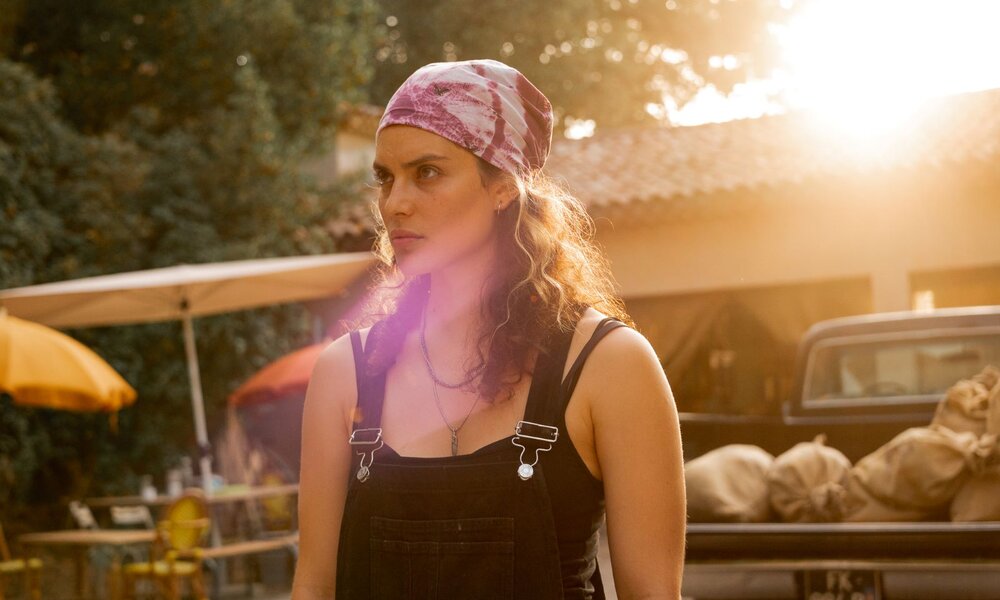 Junge Frau mit lockigem Haar und pinkem Tuch blickt ernst in die Ferne, hinter ihr Sonnenschirme und ein Pick-up im Abendlicht. | © Netflix