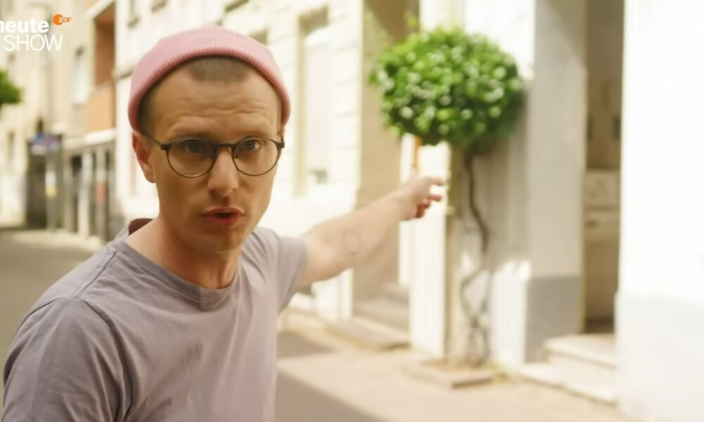 Ein junger Mann mit Brille und rosa Mütze zeigt ernst blickend auf eine helle Hausfassade mit rund geschnittenem Baum. | © YouTube / ZDF / heute-show