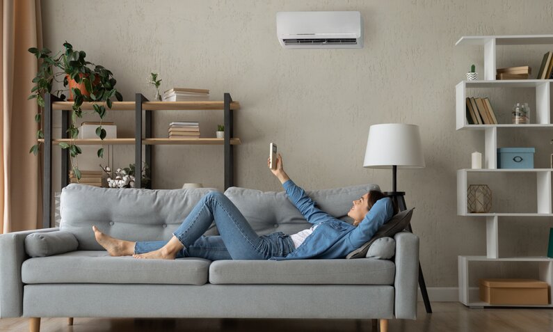 Frau liegt auf Sofa und schaltet Klimaanlage ein | © Getty Images / fizkes