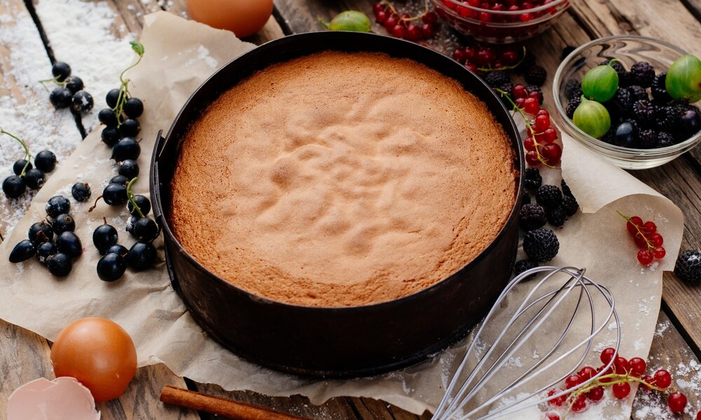 Ein fertig gebackener Biskuitboden in der Backform steht auf einem Holztisch. Unter der Kuchenform ist etwas Backpapier ausgelegt. Neben dem Kuchen befinden sich aufgeschlagene Eierschalen, Beeren und ein Schneebesen.  | © Getty Images / VladimirYa
