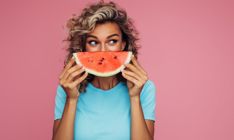 Eine junge Frau mit blonden Locken hält sich ein Stück Wassermelone vor das Gesicht. Der Hintergrund ist komplett pink eingefärbt und die Frau trägt ein hellblaues T-Shirt. | © Getty Images / CoffeeAndMilk