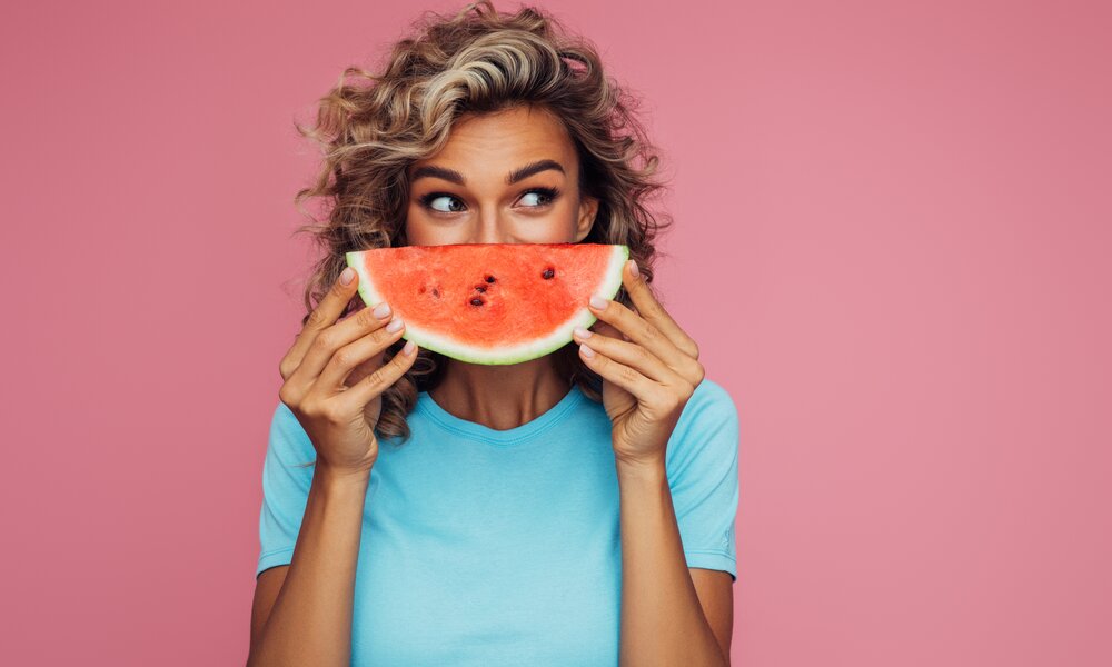 Eine junge Frau mit blonden Locken hält sich ein Stück Wassermelone vor das Gesicht. Der Hintergrund ist komplett pink eingefärbt und die Frau trägt ein hellblaues T-Shirt. | © Getty Images / CoffeeAndMilk