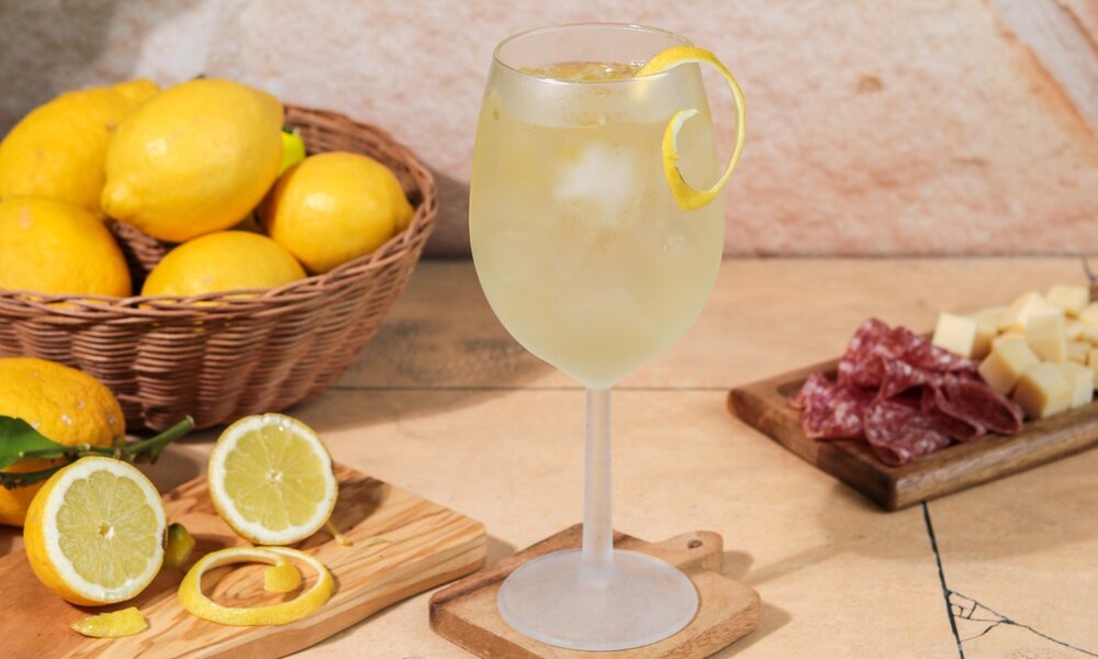 Ein Weinglas gefüllt mit Limoncello Spritz steht auf einem Holzbrett. Daneben steht ein Korb voller frischer Zitronen und eine aufgeschnittene Zitrone. Auf der anderen Seite steht ein Brett mir Salami und Käsewürfeln. | © Getty Images / MelanieMaya
