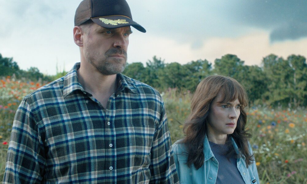 David Harbour als Jim Hopper and Winona Ryder als Joyce Byers in der Netflix Serie "Stranger Things". Die beiden stehen unter einem wolkigen Himmel auf einer Blumenwiese und sehen beide grimmig entschlossen aus.  | © Netflix