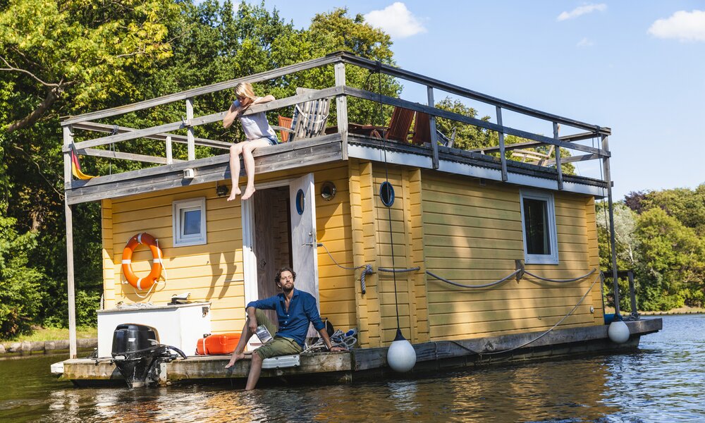 Ein Pärchen schippert mit einem gelben Hausboot über einen Fluss | ©  Getty Images / Westend61