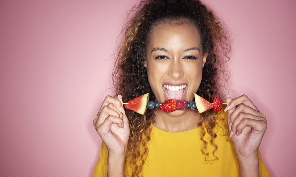 Frau steht vor rosa Hintergrund und hält einen Obst-Spieß in den Händen. Aufgespießt sind Wassermelone, Blaubeeren, Erdbeeren und Himbeeren. Die Frau hat lockiges Haar und ein trägt ein gelber T-Shirt. Sie schaut mit geöffnetem Mund frech in die Kamera. | © Getty Images / Tara Moore