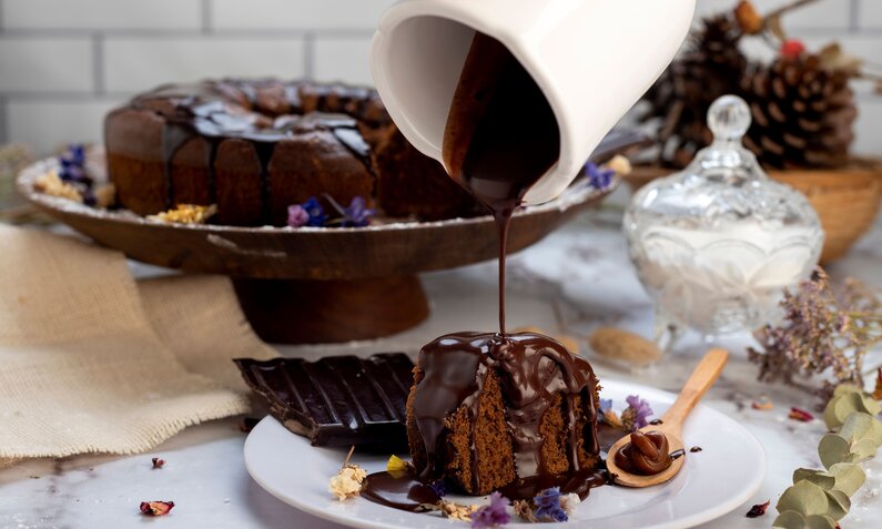 Schokokuchen auf einer Kuchenplatte, im Vordergrund ein Stück Schokokuchen, der von einer Hand mit flüssiger Schokolade überzogen wird.  | © Getty Images / Wirestock