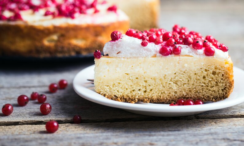 Ein Stück Johannisbeer-Torte liegt auf einem weißen Teller. Im Hintergrund und etwas unscharf sieht man die ganze Torte. | © Getty Images / ollinka