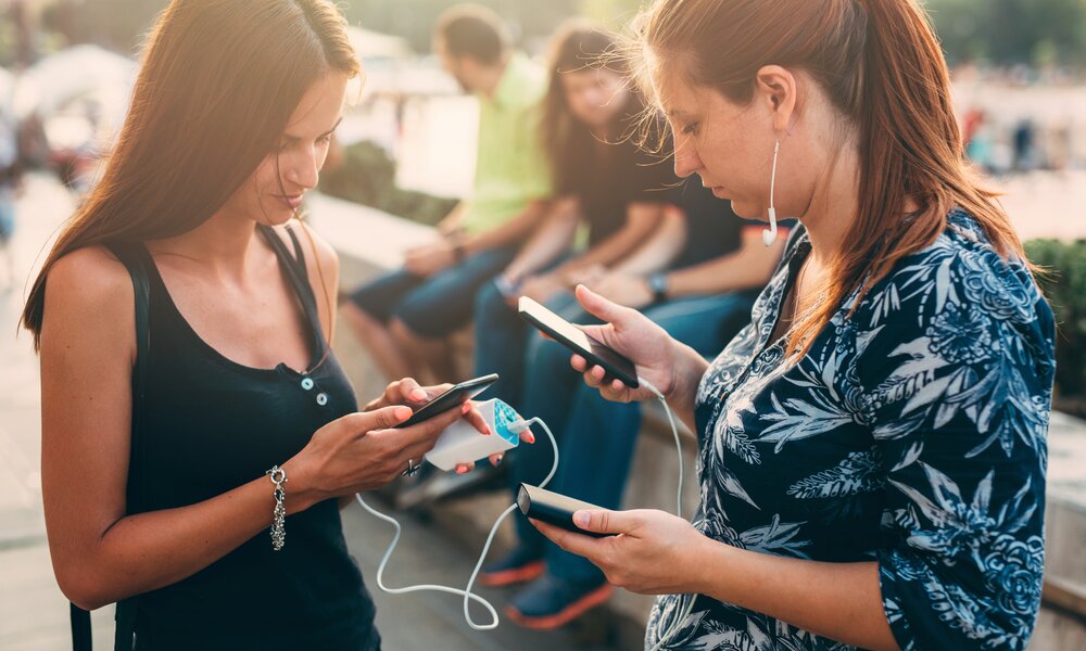 Zwei junge Frauen haben eine Power Bank an ihre handys angeschlossen und schauen auf ihr jeweiliges Display. Die Geräte sind mit weißen Kabeln verbunden. | © Getty Images/Todor Tsvetkov