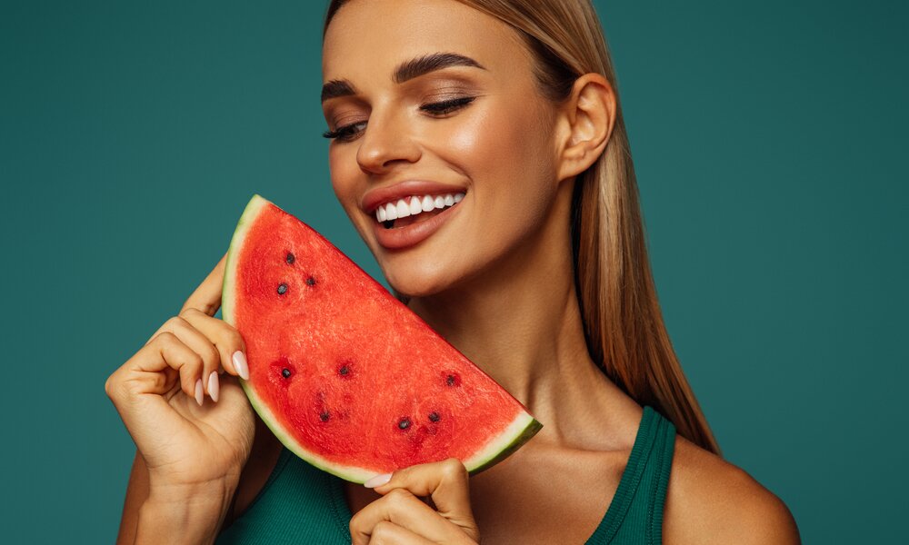 Junge Frau mit blonden Haaren hält ein Stück Wassermelone in den Händen. Sie schaut auf die Melone und lächelt dabei. Der Hintergrund ist türkis unterlegt. | © Getty Images / CoffeeAndMilk