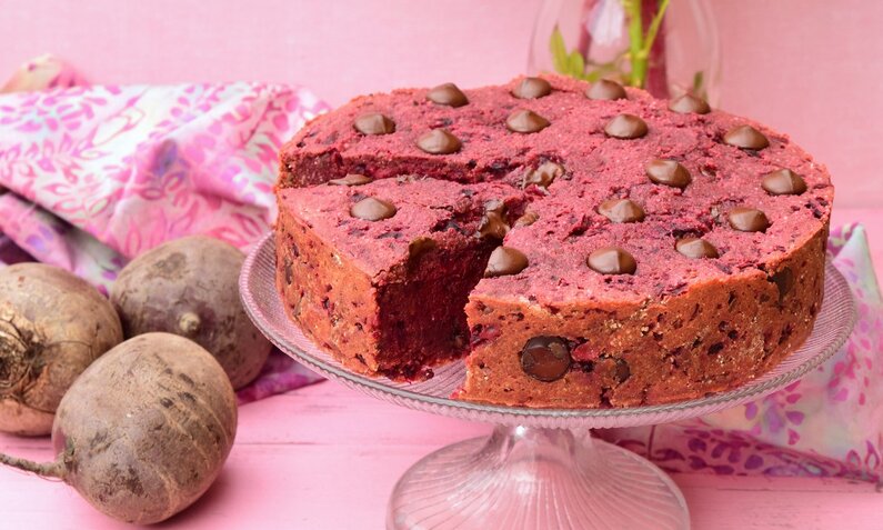 Rote-Bete-Kuchen steht auf einem Kuchenteller. Der Hinterggrund ist rosa und neben dem Kuchen liegt frische Rote-Bete. | © Getty Images / AmalliaEka