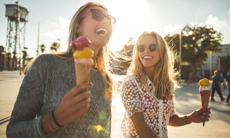 Zwei junge Frauen in Nahaufnahme lachen sich an und tragen Sonnenbrillen. Jede hält ein Eis in der Hand. Es scheint die Sonne und es herrscht ein sommerliches Ambiente.  | © Getty Images / Georgijevic