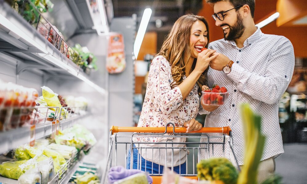 Ein Mann und eine Frau sind im Supermarkt. Sie schieben einen Einkaufswagen und der Mann reicht der Frau eine Schale mit Erdbeeren. Die Frau probiert eine Erdbeere und beide lächeln dabei.  | © Getty Images / Aja Koska