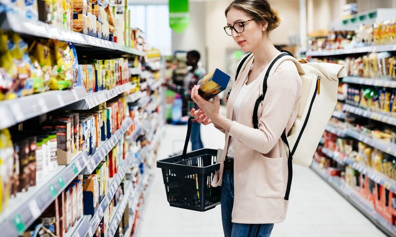 Frau mit Brille und geschlossenen Haaren steht im Supermarkt von einem Regal mit einem Produkt in der Hand. Sie trägt außerdem einen Einkaufskorb und einen Rucksack.  | © Getty Images / Tom Werner
