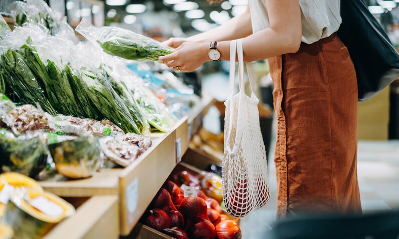 Frauenkörper steht vor einem Gemüse- und Obstregal im Supermarkt. Sie trägt einen Stoffbeutel mit Löchern und greift mit der Hand die Auslage. | © Getty Images / d3sign