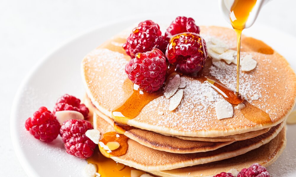 Pancakes getoppt mit Puderzucker und Himbeeren auf einem weißen Teller. Der Teller steht auf einem weißen Untergrund und es fließt Ahornsirup aus einer Sauciere auf die Pancakes. | © Getty Images / vaaseenaa
