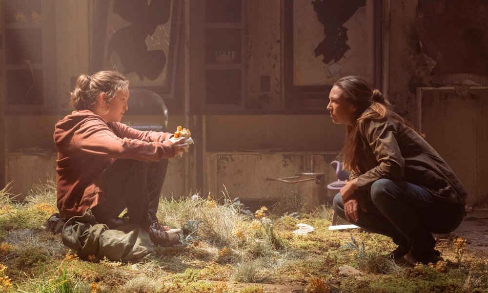 Ellie-Darstellerin Belly Ramsay sitzt in einem Lichtstrahl auf dem Boden eines Raums, der mit Grün bewachsen ist. Tess-Darstellerin Anna Torv hockt rechts von ihr und spricht mir ihr. | © HBO
