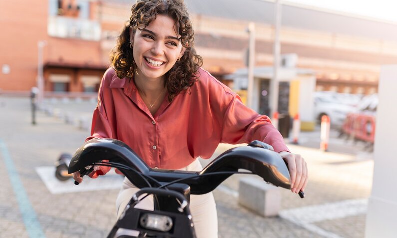 Eine junge Frau lächelt über den Lenker ihres E-Bikes hinweg. Sie trägt eine rote Bluse und eine weiße Hose und ist brünett.