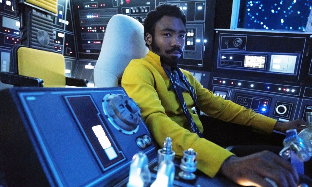 Donald Glover als Lando Calrissian im Kinofilm "Solo: A Star Wars Story". Er sitzt im Pilotensessel eines kleinen Raumschiffs in einem gelben Suit in bläuöichem Licht. | © ddp images/Jonathan Olley /© Lucasfilm/ © Walt Disney Studios Motion Pictures /Courtesy Everett Collection
