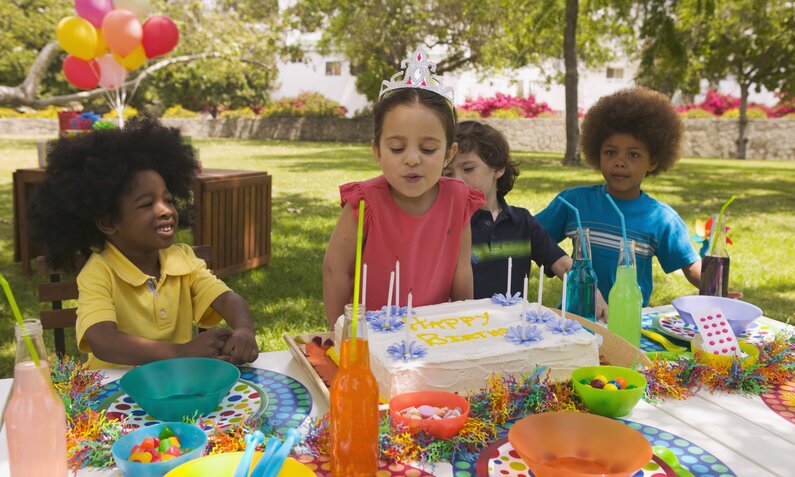 Viele Kinder sitzen draußen an einem bunten Geburtstagstisch. Ein Mädchen pustet Kerzen auf dem Kuchen aus. | © Getty Images / Burke/Triolo Productions