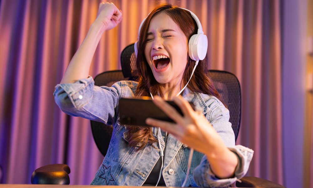 Junge asiatische Frau jubelt, während sie an einem Tisch sitzt und mit ihrem Smartphone spielt. Sie trägt ein Paar weiße Kopfhörer dabei. | © Getty Images/jackieenjoyphotography