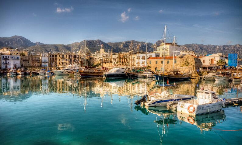 Der Hafen der Stadt Girne auf Zypern mit Fischerbooten vor einer Kulisse von alten Häusern, dahinter ein Bergpanorama | © Getty Images / nejdetduzen