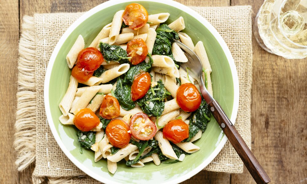 Teller gefüllt mit Penne, Spinat und Cherrytomaten. Der Teller steht auf einem beigen Platzdeckchen. | © Getty Images / Westend61