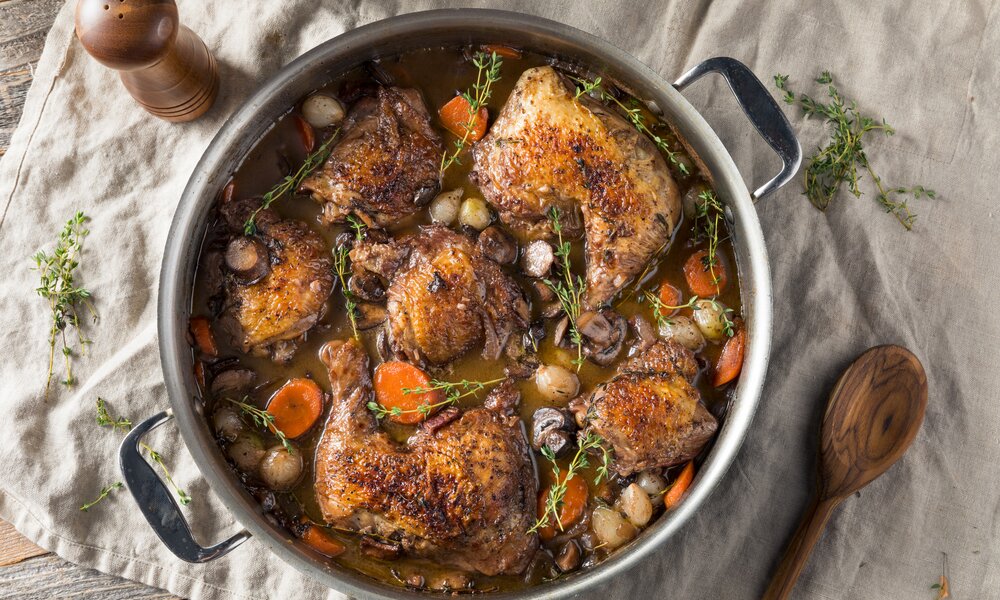 Hühnerschenkel a la Coq au Vin in einem runden Bräter zusammen mit frischen Kräutern, Kartoffeln und Möhren. | © Getty Images / bhofack2