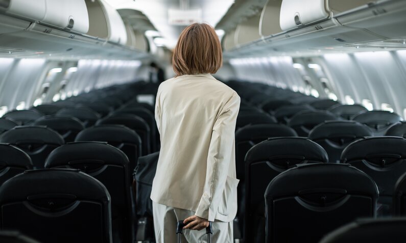 Frau geht mit ihrem Handgepäckskoffer durch ein Flugzeug, die anderen Sitze sind leer | ©  Getty Images / Susumu Yoshioka