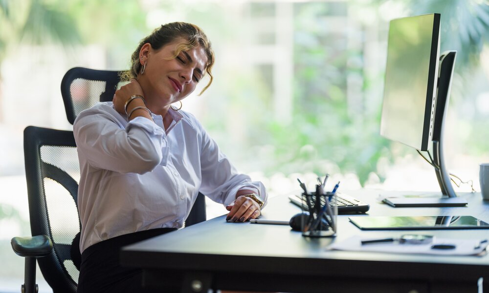 Frau sitzt am Computer im Büro und hält sich mit einem schmerzverzerrten Gesicht den Nacken  | © Getty Images / ilkercelik