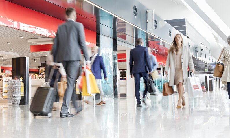  Leute mit Koffern laufen durch den Flughafen, im Hintergrund sind Shops zu sehen  | ©  Getty Images / Caia Image
