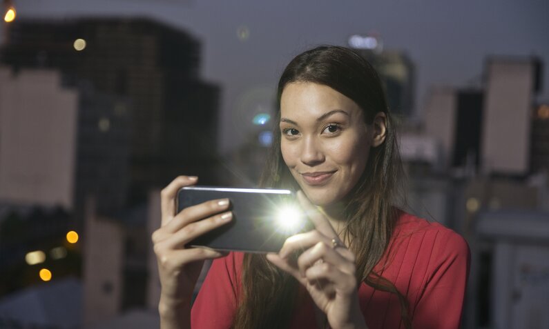 EIne junge Frau lächelt in die kamera, während sie in der Hand ein Smartphone mit eingeschalteter Taschenlampenfunktion hält. Im Hintergrund sind die Silhouetten von Häusern zu erkennen. | © Getty Images/Westend61