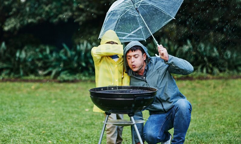 Mann steht bei Regen vorm Grill, ein Kind hält ihm einen Regenschirm  | ©  Getty Images / Adene Sanchez