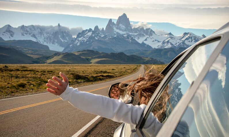 Frau lehnt sich aus dem Autofenster, im Hintergrund eine Bergkulisse | ©  Getty Images / Paul Biris