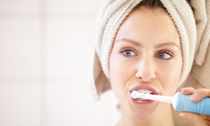 Eine junge Frau mit einem Handtuch auf dem Kopf putzt sich mit einer elektrischen Zahnbürste die Zähne. | © Getty Images/PeopleImages