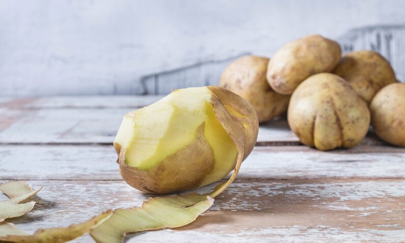 Eine halbgeschälte Kartoffel, im Hintergrund ein Haufen ungeschälter Kartoffeln | © Getty Images / Caterina Oltean / 500px