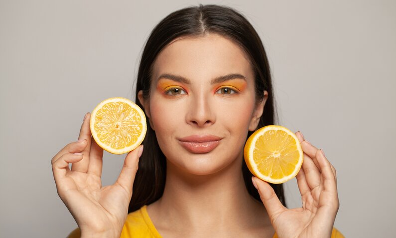 Eine junge Frau mit dunklen Haaren und orangem Makeup hält zwei Orangenschalen in den Händen. | © Getty Images / JNemchinova